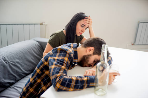 Concerned woman sitting beside a man affected by alcohol misuse, with an empty bottle on the table in a home setting.