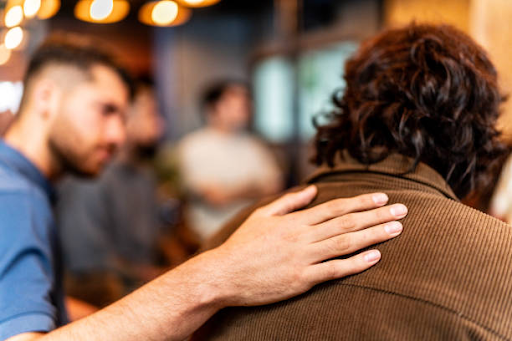 Person offering reassurance by placing a supportive hand on another person's shoulder during an addiction recovery support group meeting.