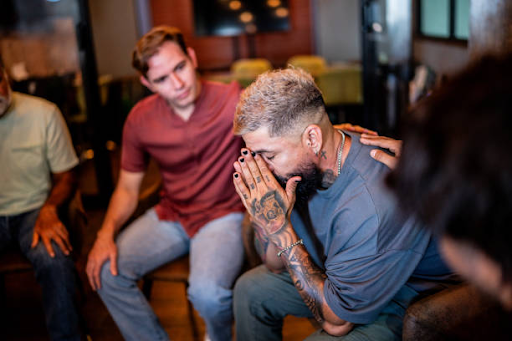 Person receiving emotional support during a group addiction recovery meeting, with another participant offering comfort by placing a hand on their shoulder.