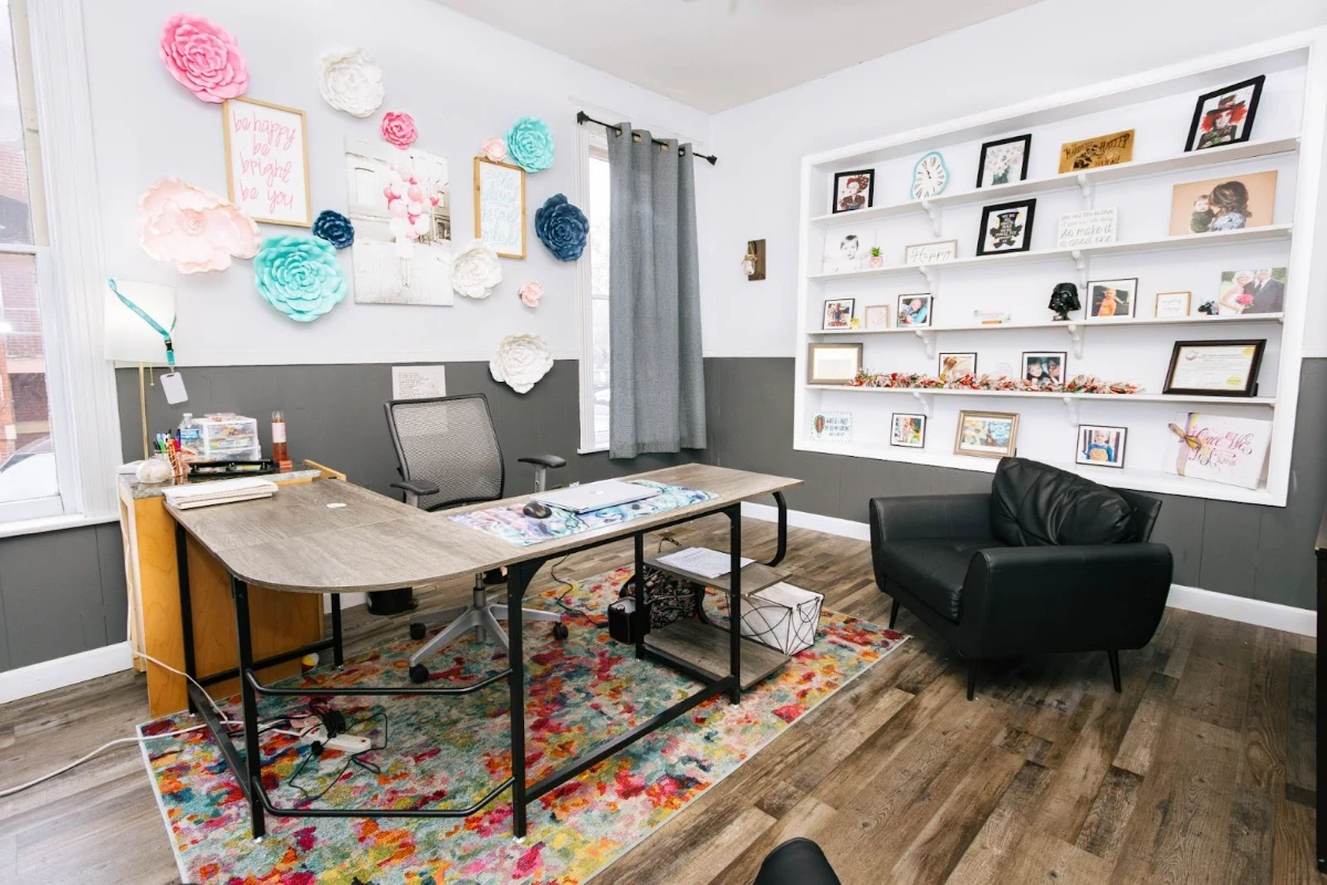 A bright and welcoming therapy office interior featuring an L-shaped wooden desk, and white shelving displaying photos.