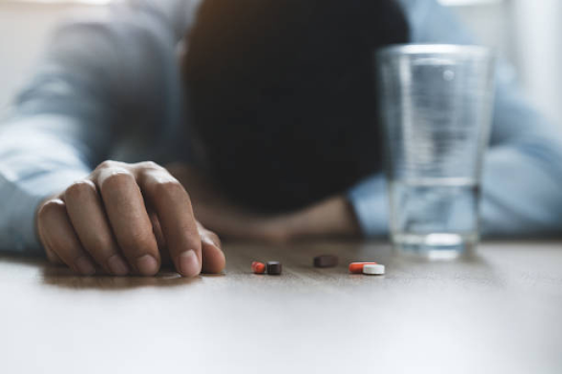 Man resting his head on a table beside scattered pills and a glass of water