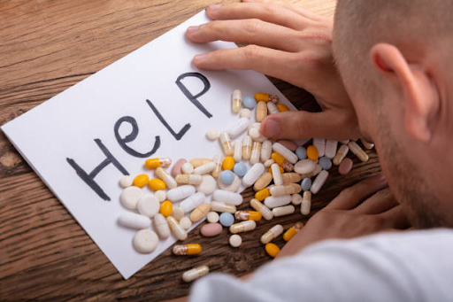 Man reaching toward scattered pills on a table beside a paper with the word &ldquo;Help&rdquo; written on it