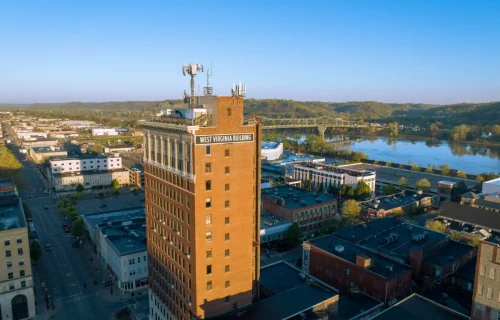Aerial view of the West Virginia Building towering over a downtown riverfront city under a clear blue sky.