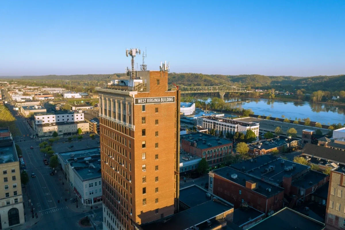 Aerial view of the West Virginia Building towering over a downtown riverfront city under a clear blue sky.