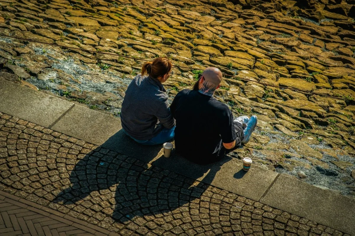 Two people sitting together on stone steps outdoors, seen from above, each with a takeaway coffee cup beside them.