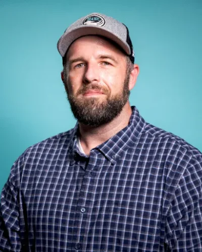 Joshua Carter, Behavioral Health Technician at HAWC Recovery, wearing a plaid shirt and cap against a teal background.