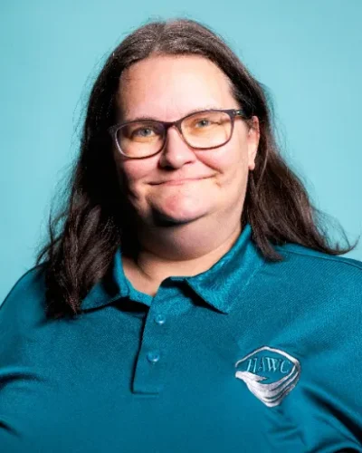 Pamela Roberts, counselor at HAWC Recovery, smiling in teal polo shirt with HAWC logo, against a light blue background, reflecting the supportive and compassionate environment of the team.