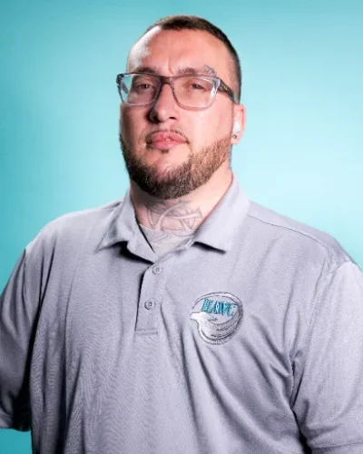 Zachery Hodge, Supportive Counselor at HAWC Recovery, wearing a gray polo shirt with the HAWC logo, posing against a turquoise background.