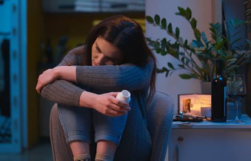Woman sitting alone holding a pill bottle with her head resting on her arms indoors at night