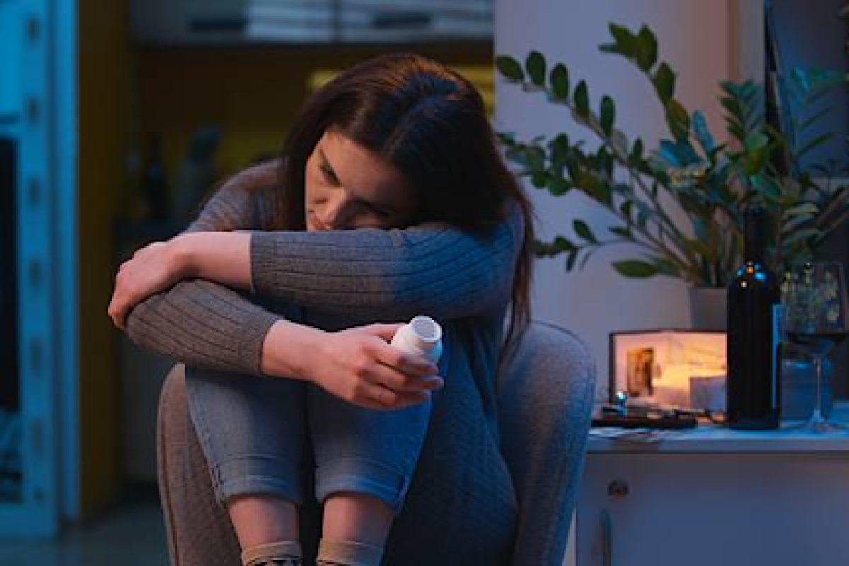 Woman sitting alone holding a pill bottle with her head resting on her arms indoors at night