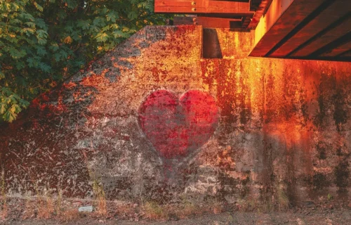 A faded red heart painted as graffiti on a weathered concrete wall beneath a bridge, surrounded by green trees.