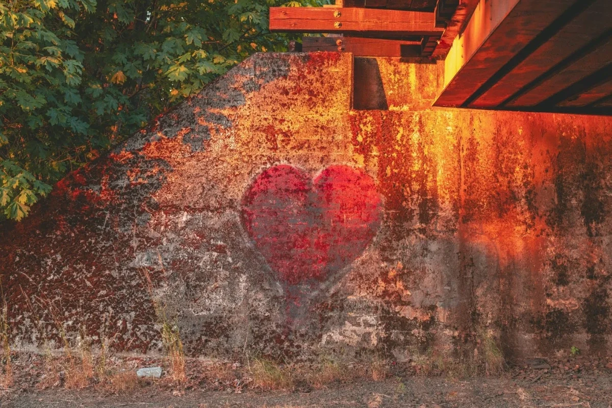 A faded red heart painted as graffiti on a weathered concrete wall beneath a bridge, surrounded by green trees.