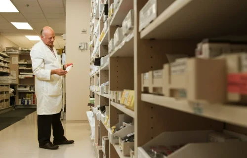 A pharmacist examining a medication package while standing between tall shelves stocked with pharmaceutical supplies.