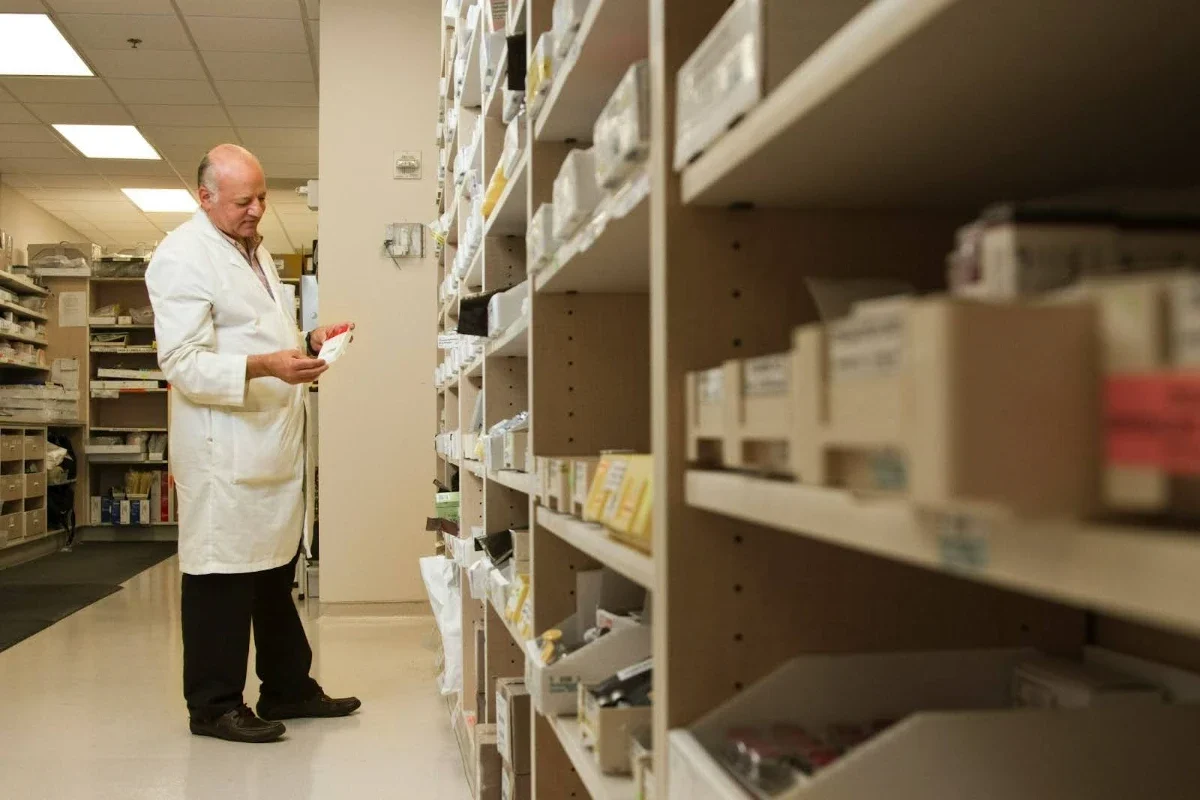 A pharmacist examining a medication package while standing between tall shelves stocked with pharmaceutical supplies.
