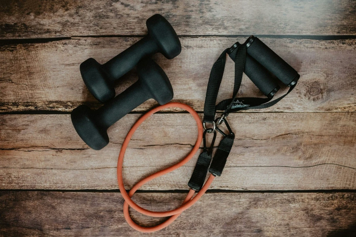 Black dumbbells and an orange resistance band with black handles placed on a rustic wooden surface.
