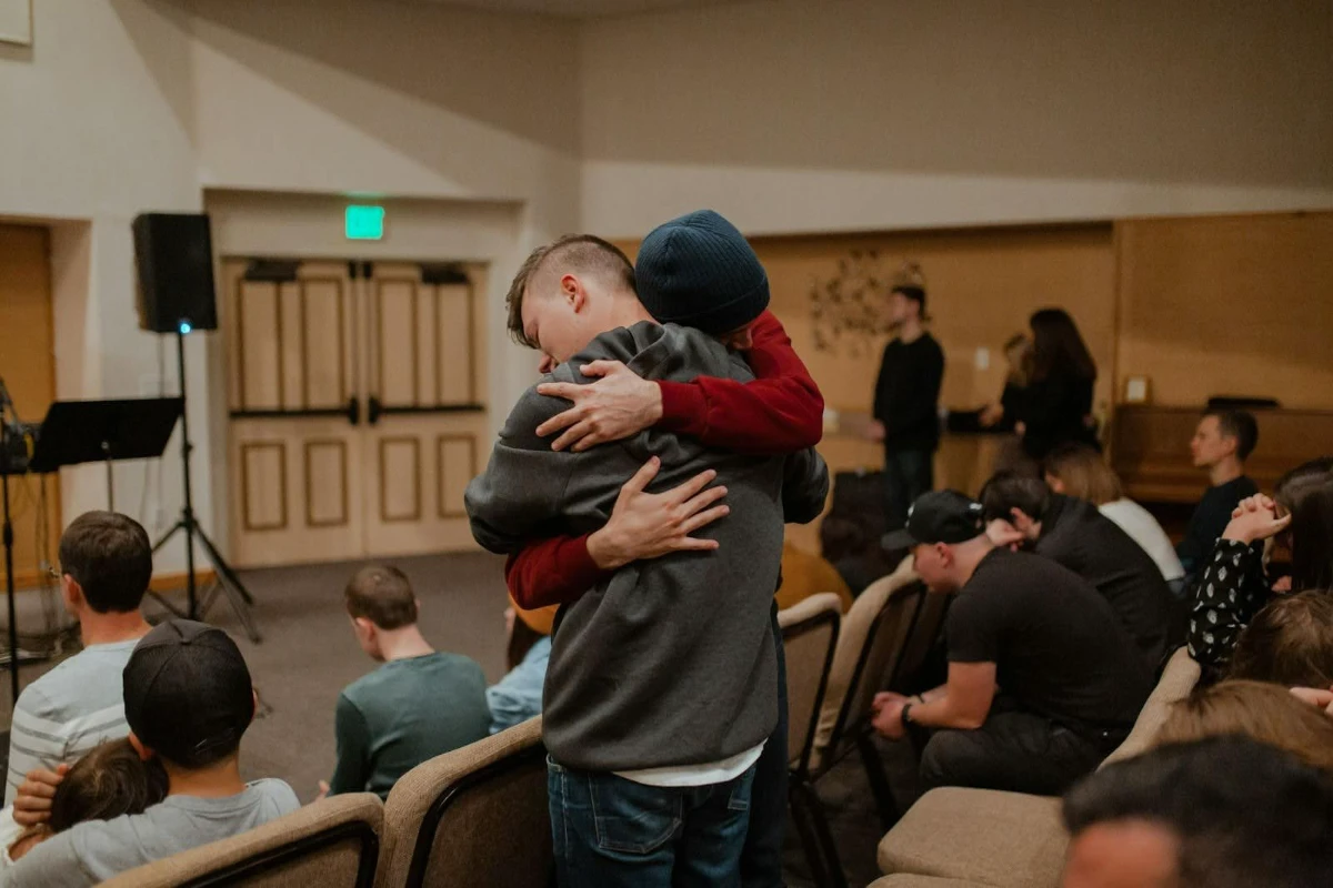 Two people sharing a warm embrace in the aisle of a community gathering hall, with other attendees seated around them.